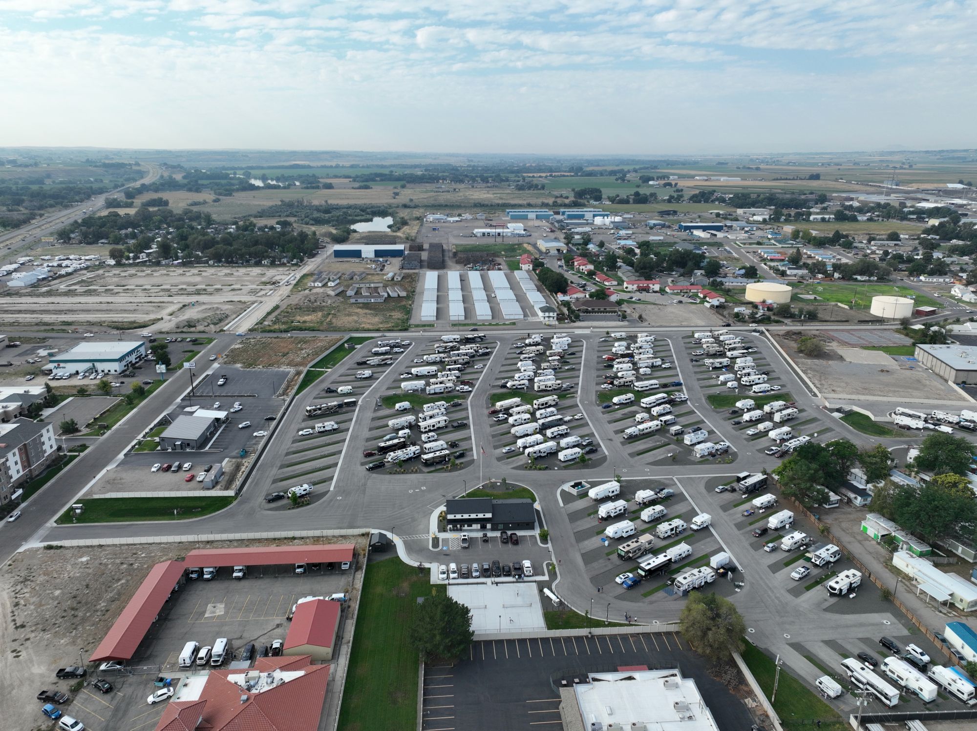 An aerial view of a busy car dealership lot with rows of vehicles, surrounding buildings, and open fields in the distance, under a partly cloudy sky.