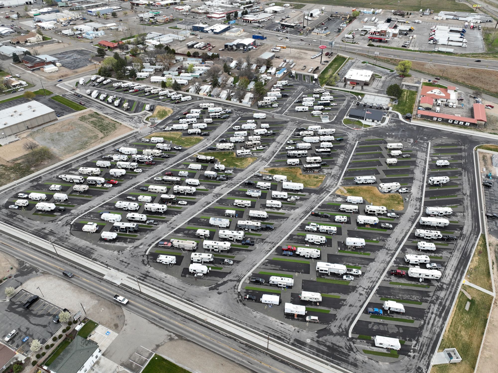 An aerial view of a large RV park or campground with numerous white trailers and vehicles arranged in rows, near roads and some buildings, on a paved lot.