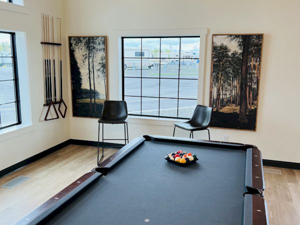 A game room with a pool table, black felt, a fruit bowl on the table, and triptych forest wall art beside a large window, chairs around the room.