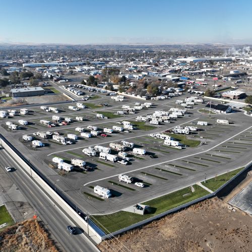 An aerial view of a large RV park with rows of campers and mobile homes spread across grassy lots, bordered by roads and distant buildings, under a clear sky.