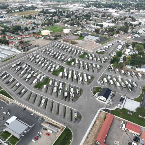 Aerial view of a car lot with many white vehicles parked in organized rows; surrounding roads, buildings, and greenery.