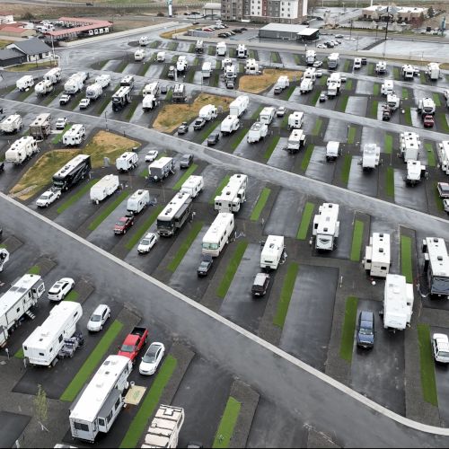 Aerial view of a large RV and trailer parking lot with rows of campers, buses, and vans arranged on paved spots and grassy strips.