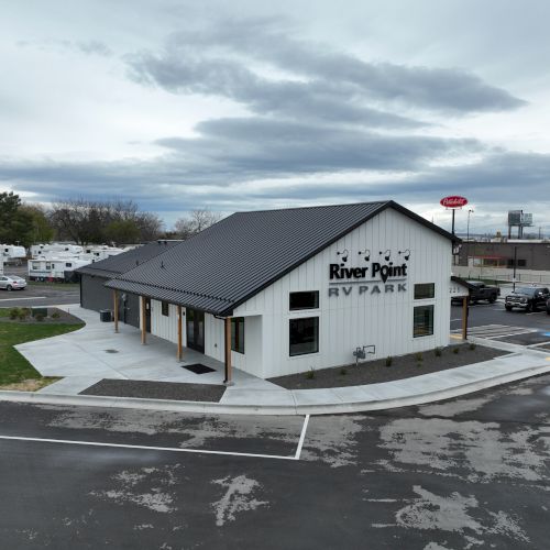 A small roadside RV park office with a gray gabled roof, sign reading &ldquo;Riv e&hellip; RV Park,&rdquo; a parking lot, and several RVs in the background.